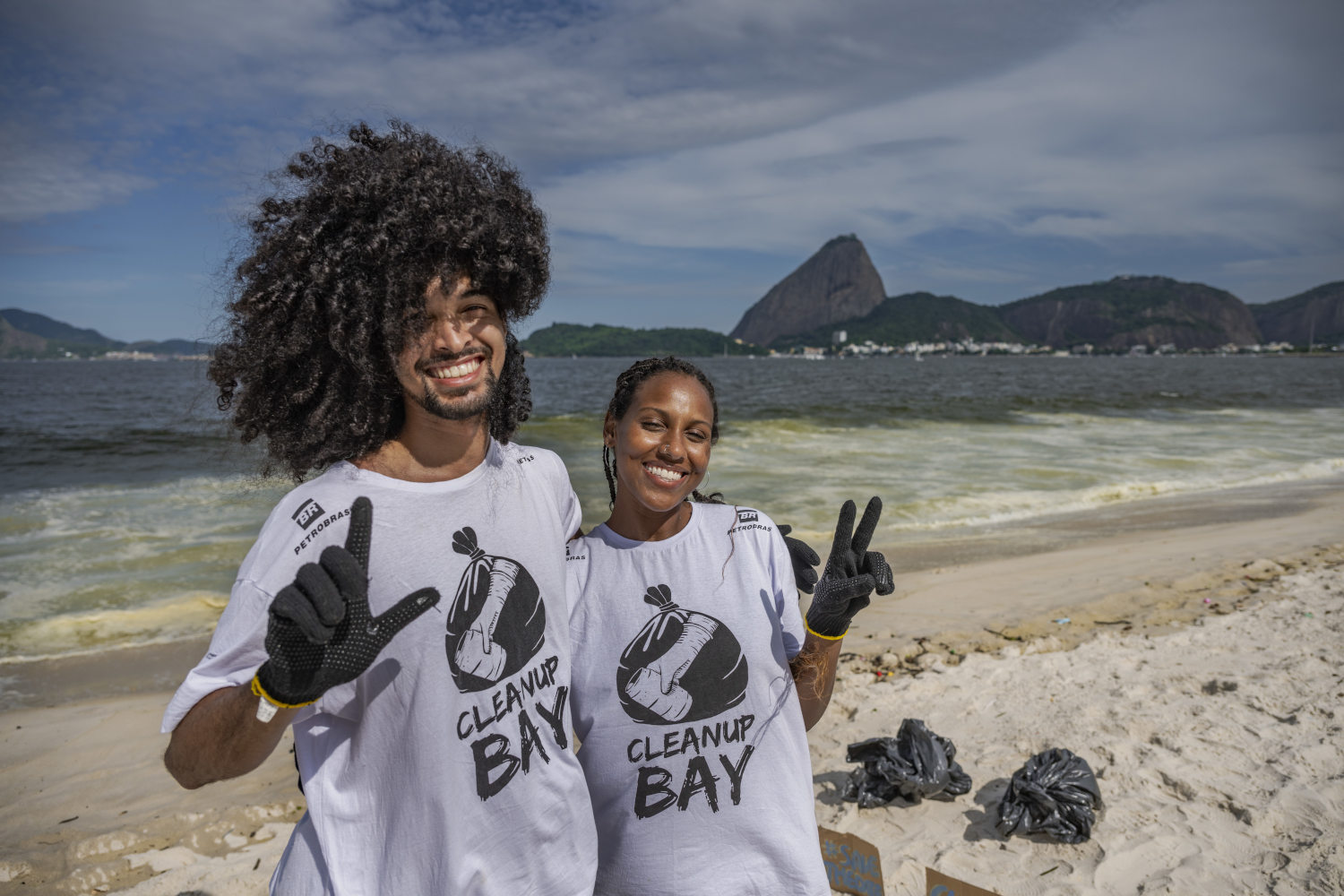 Homem e mulher posando para foto com camisa da iniciativa Clean Up Bay na praia com corcovado ao fundo